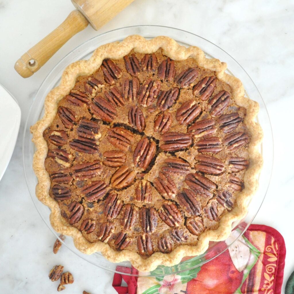 A freshly baked pecan pie in a glass pie dish, topped with whole pecans arranged in a circular pattern, sits on a marble countertop next to a rolling pin and a festive red cloth.