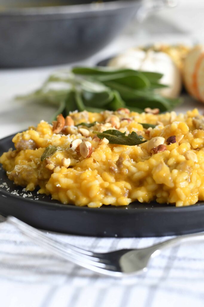 A close-up of creamy risotto with pumpkin and nuts, garnished with fresh sage, served on a black plate. A fork rests nearby, and blurred ingredients are visible in the background.