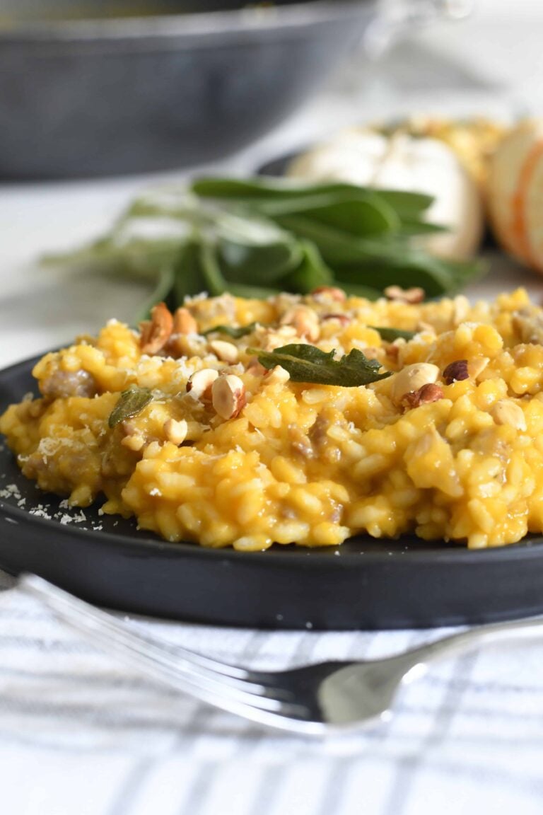 A close-up of creamy risotto with pumpkin and nuts, garnished with fresh sage, served on a black plate. A fork rests nearby, and blurred ingredients are visible in the background.