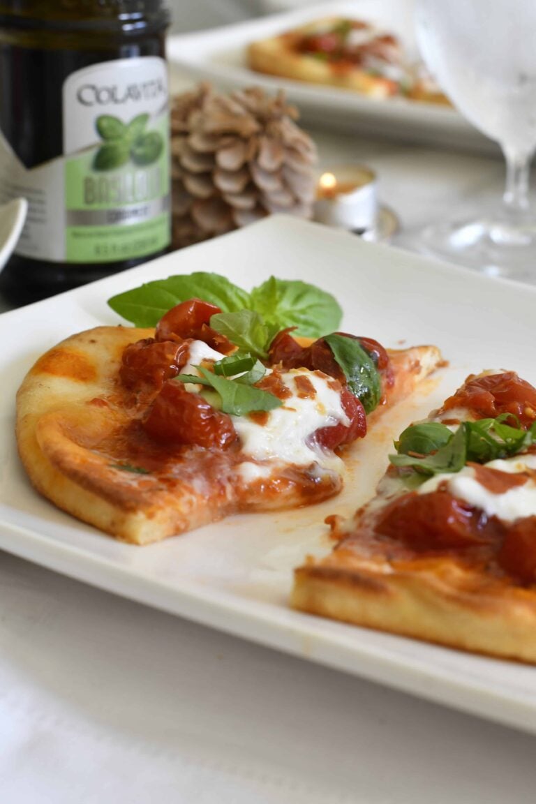 A close-up of a plate with two slices of margherita pizza topped with melted cheese, cherry tomatoes, and fresh basil, set on a white table next to a basil oil bottle, pinecone, and glass of water.
