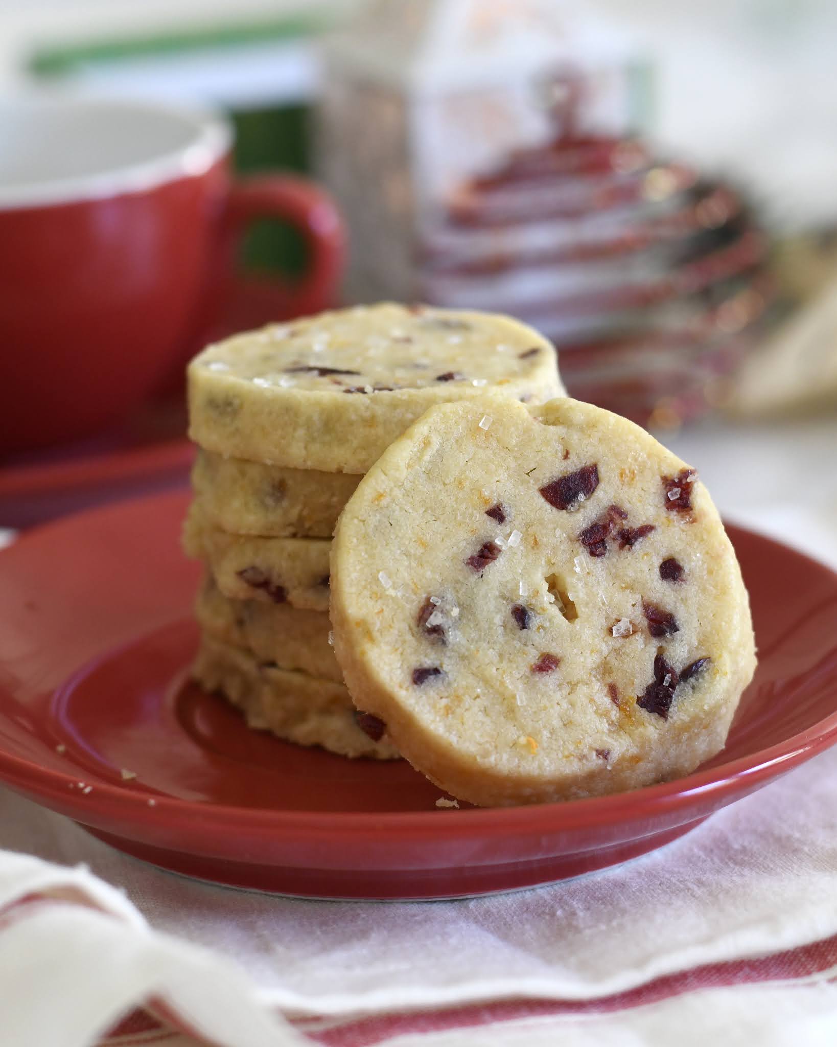 A stack of round, shortbread cookies with visible bits of dried cranberries sits on a red plate, with a red cup and holiday decorations blurred in the background.