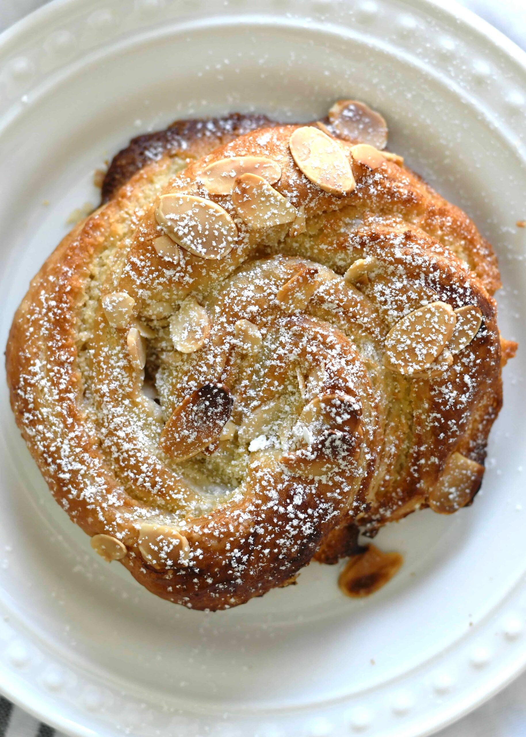 A close-up of a spiral pastry topped with sliced almonds and dusted with powdered sugar, served on a white plate.