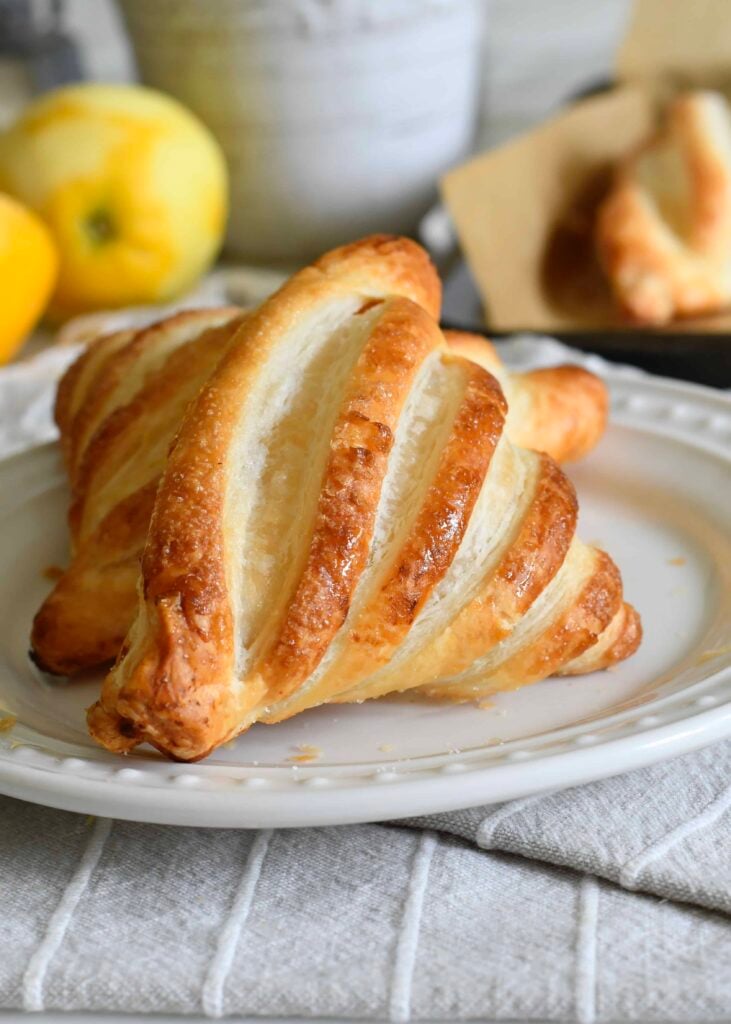 A golden, flaky pastry with slits across the top sits on a white plate, with blurred lemons and another pastry in the background.
