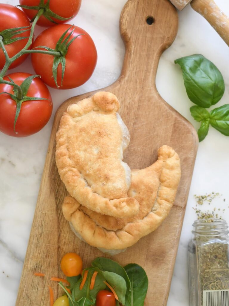 Two golden-brown calzones on a wooden cutting board, surrounded by fresh tomatoes, basil leaves, spinach, cherry tomatoes, shredded carrots, and a jar of dried herbs on a marble countertop.