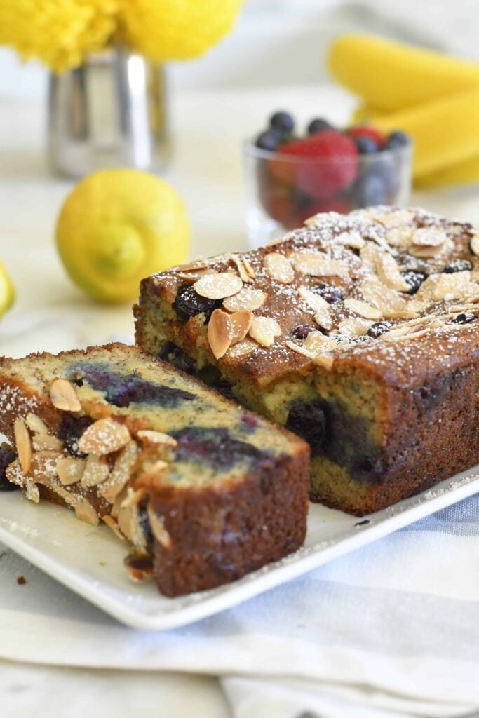 A loaf of blueberry almond cake, topped with powdered sugar and almond slices, sits on a white plate. Two slices are cut, revealing a moist texture and blueberries inside. Lemons, flowers, and berries are in the blurred background.