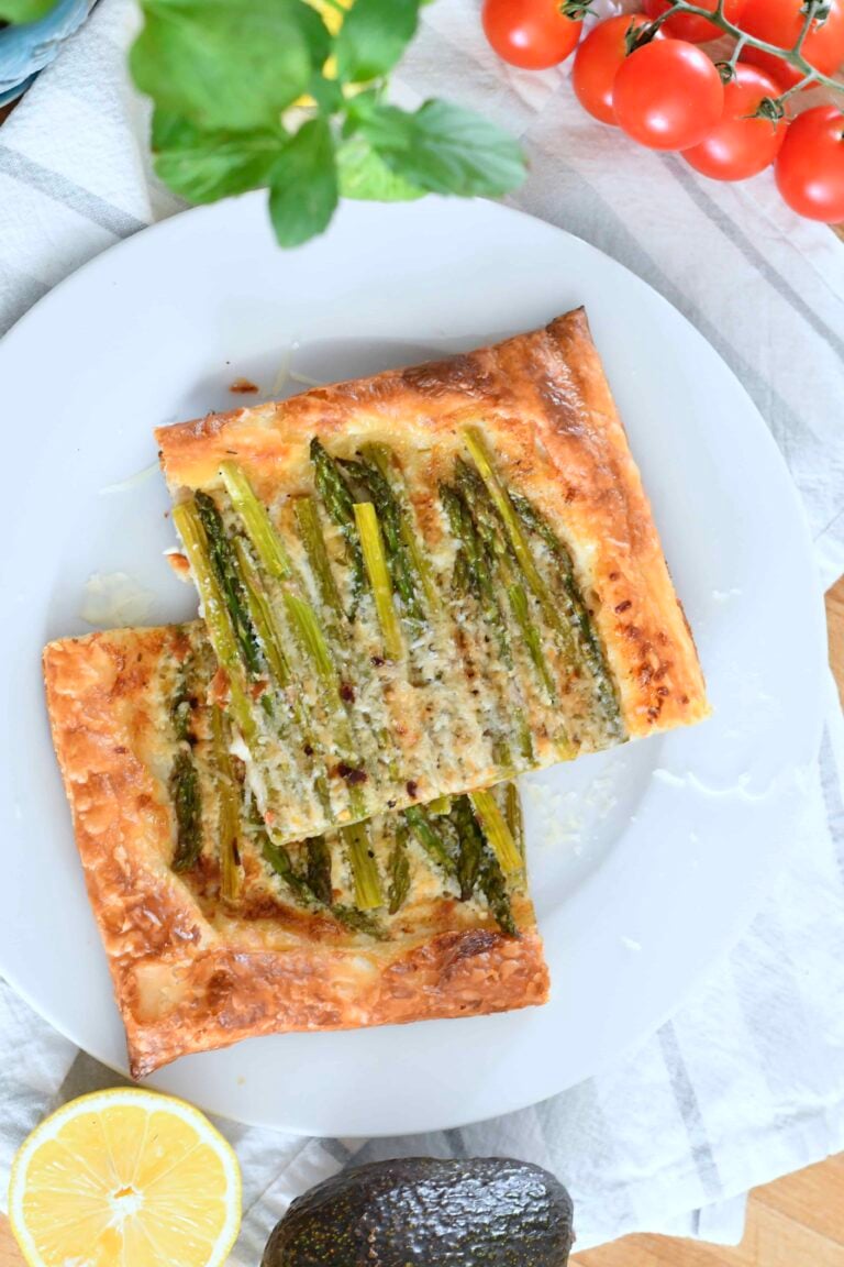A white plate holds two rectangular slices of puff pastry topped with asparagus and grated cheese. The plate is on a striped cloth next to cherry tomatoes, a halved lemon, an avocado, and fresh herbs.