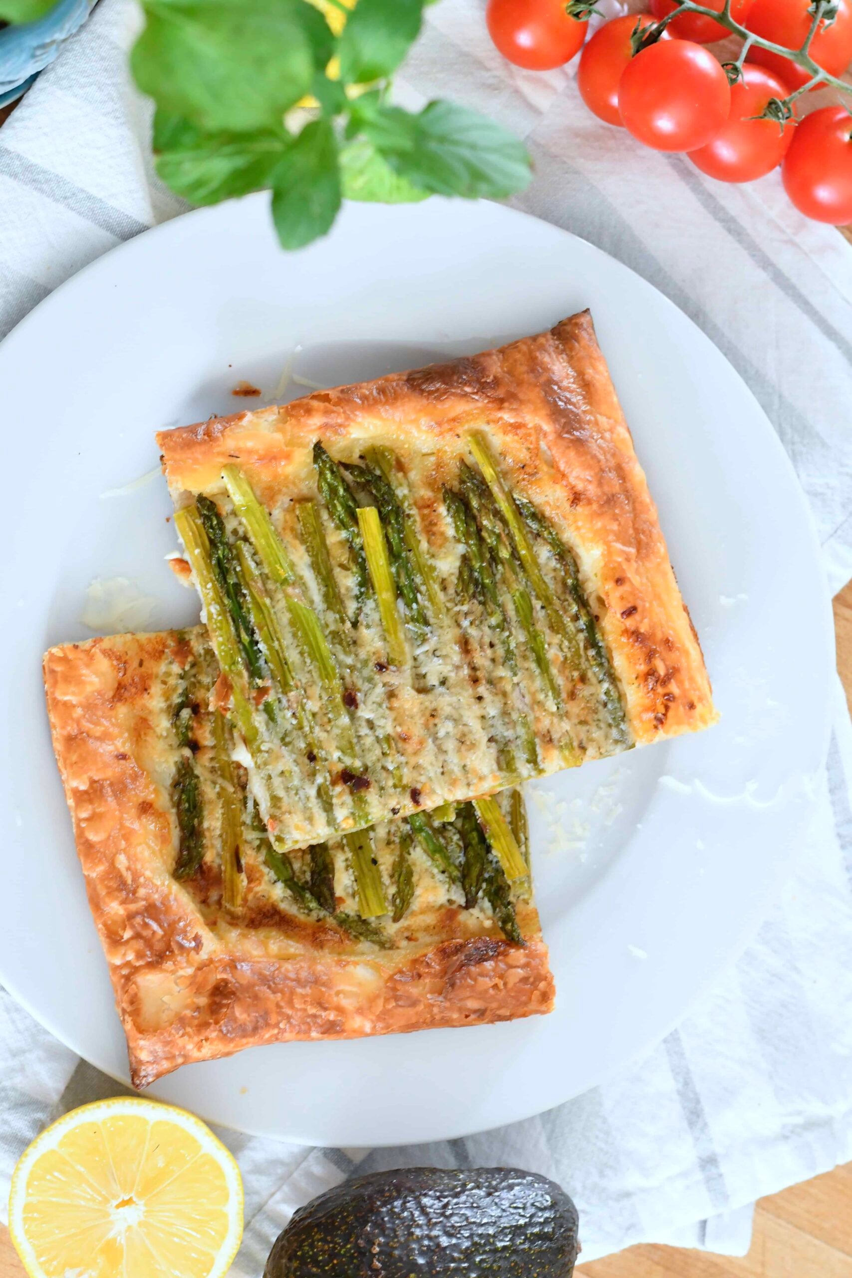 A white plate holds two rectangular slices of puff pastry topped with asparagus and grated cheese. The plate is on a striped cloth next to cherry tomatoes, a halved lemon, an avocado, and fresh herbs.