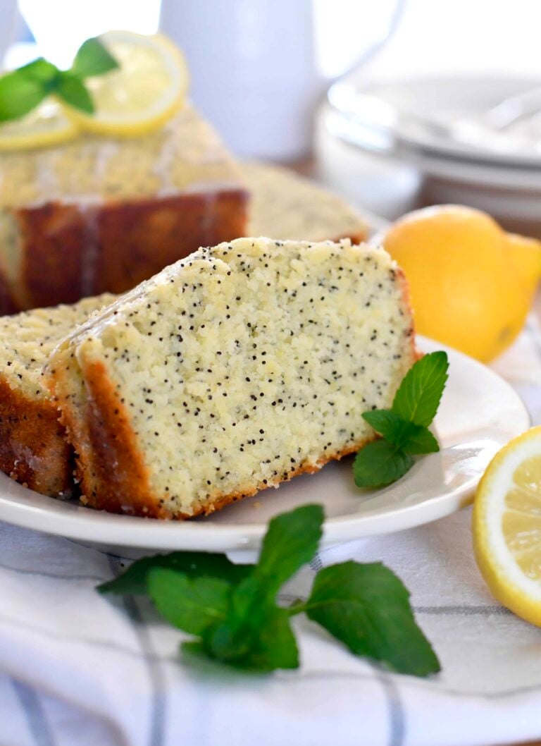 A slice of lemon poppy seed bread sits on a white plate, garnished with fresh mint and lemon slices. The bread is moist with visible poppy seeds, and a loaf and lemon are seen in the background.