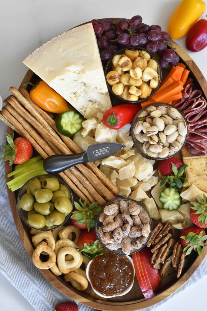A charcuterie board with assorted cheeses, crackers, breadsticks, nuts, olives, sliced vegetables, grapes, strawberries, cured meats, a spread, and a small cheese knife in the center.