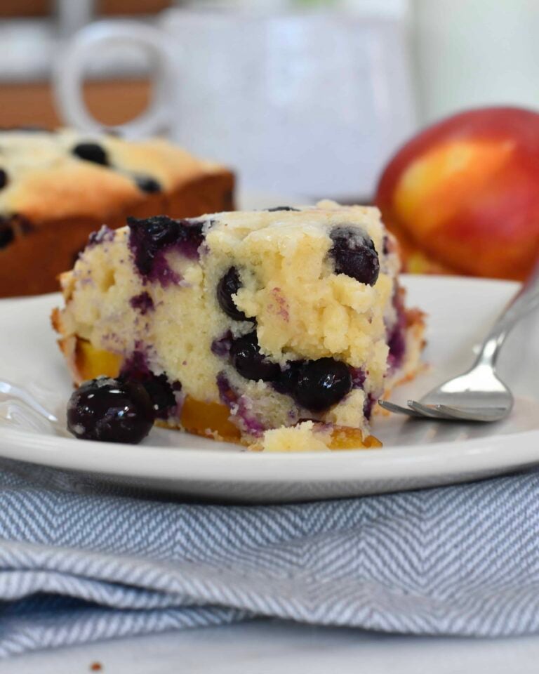 A slice of blueberry and peach cake sits on a white plate with a fork beside it. Fresh blueberries are visible in the moist cake, with a peach and the rest of the cake in the background.