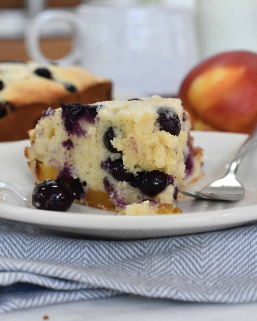 A slice of blueberry and peach cake sits on a white plate with a fork beside it. Fresh blueberries are visible in the moist cake, with a peach and the rest of the cake in the background.