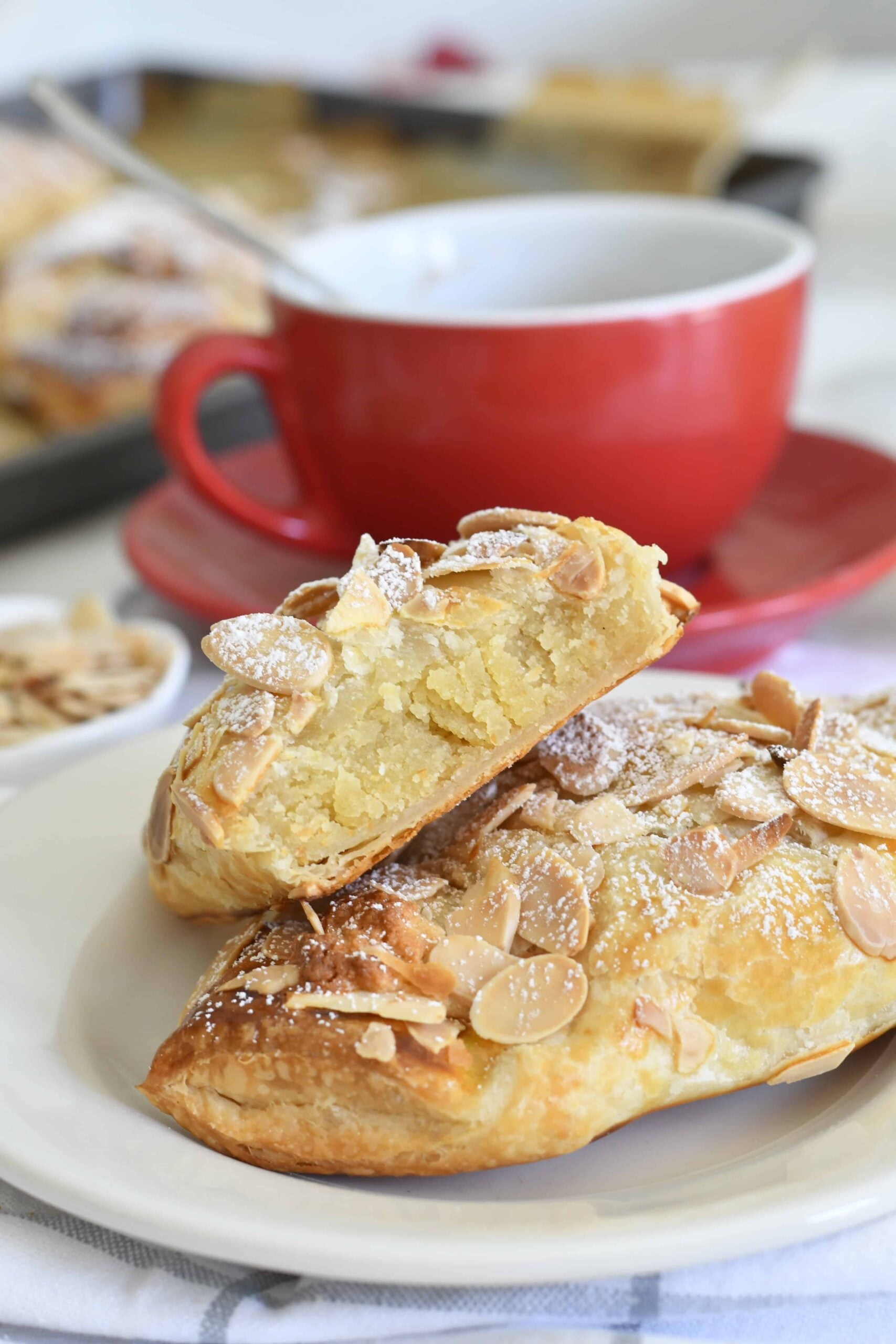 A close-up of two almond croissants dusted with powdered sugar on a white plate, with one croissant cut open to show the filling. A red cup and saucer with a hot drink are in the background.