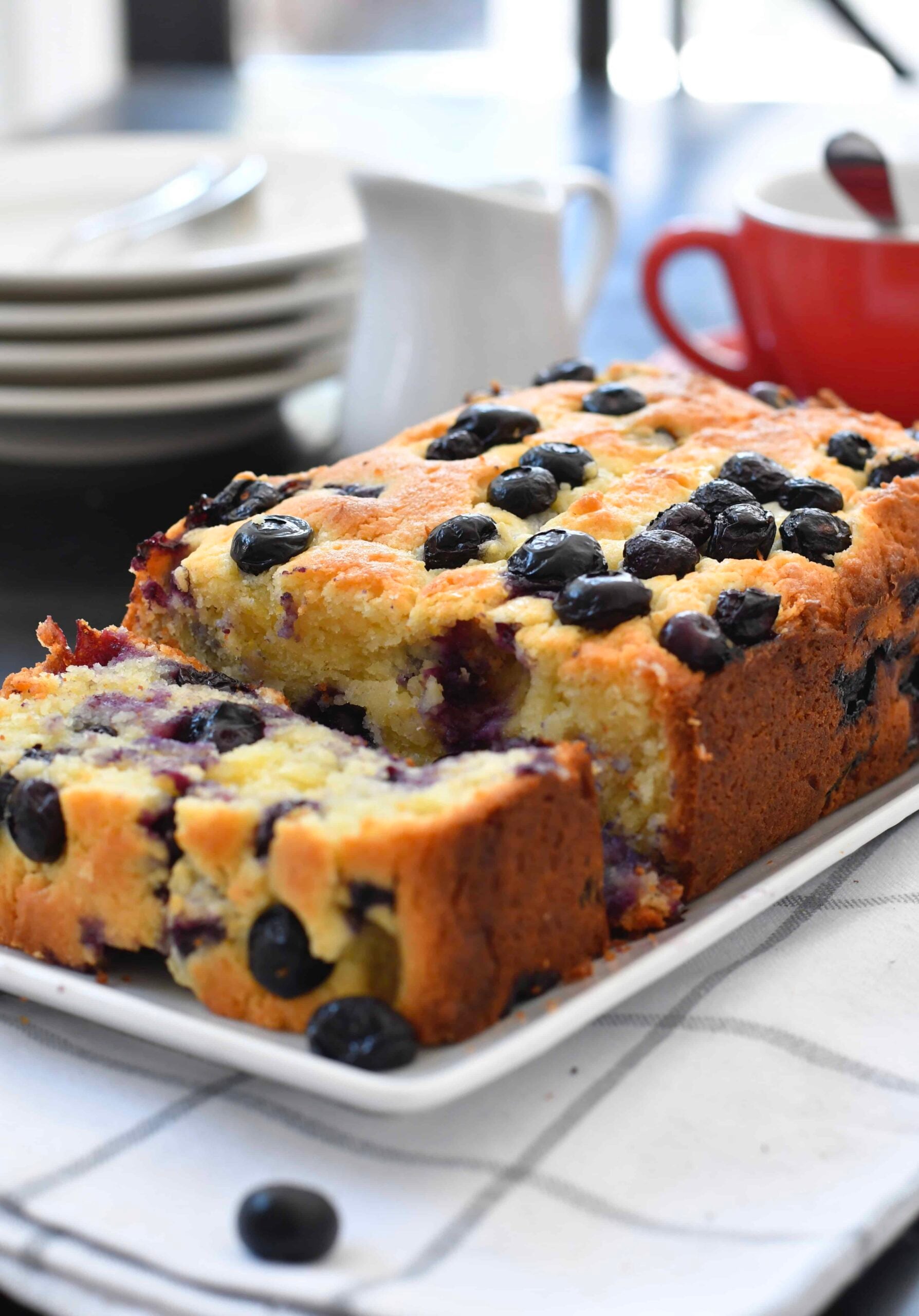 A loaf of blueberry bread on a white rectangular plate, with one thick slice cut. Fresh blueberries are visible in the bread. In the background, there are stacked plates, a white jug, and a red cup and saucer.