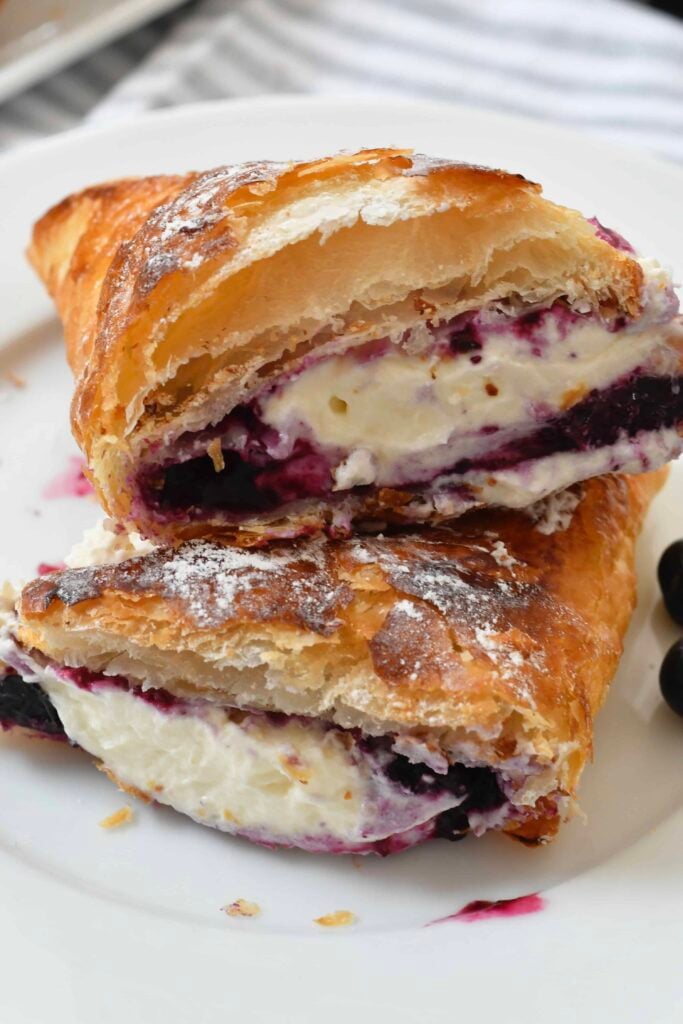 A close-up of a flaky croissant cut in half, filled with creamy cheese and blueberry filling, stacked on a white plate.