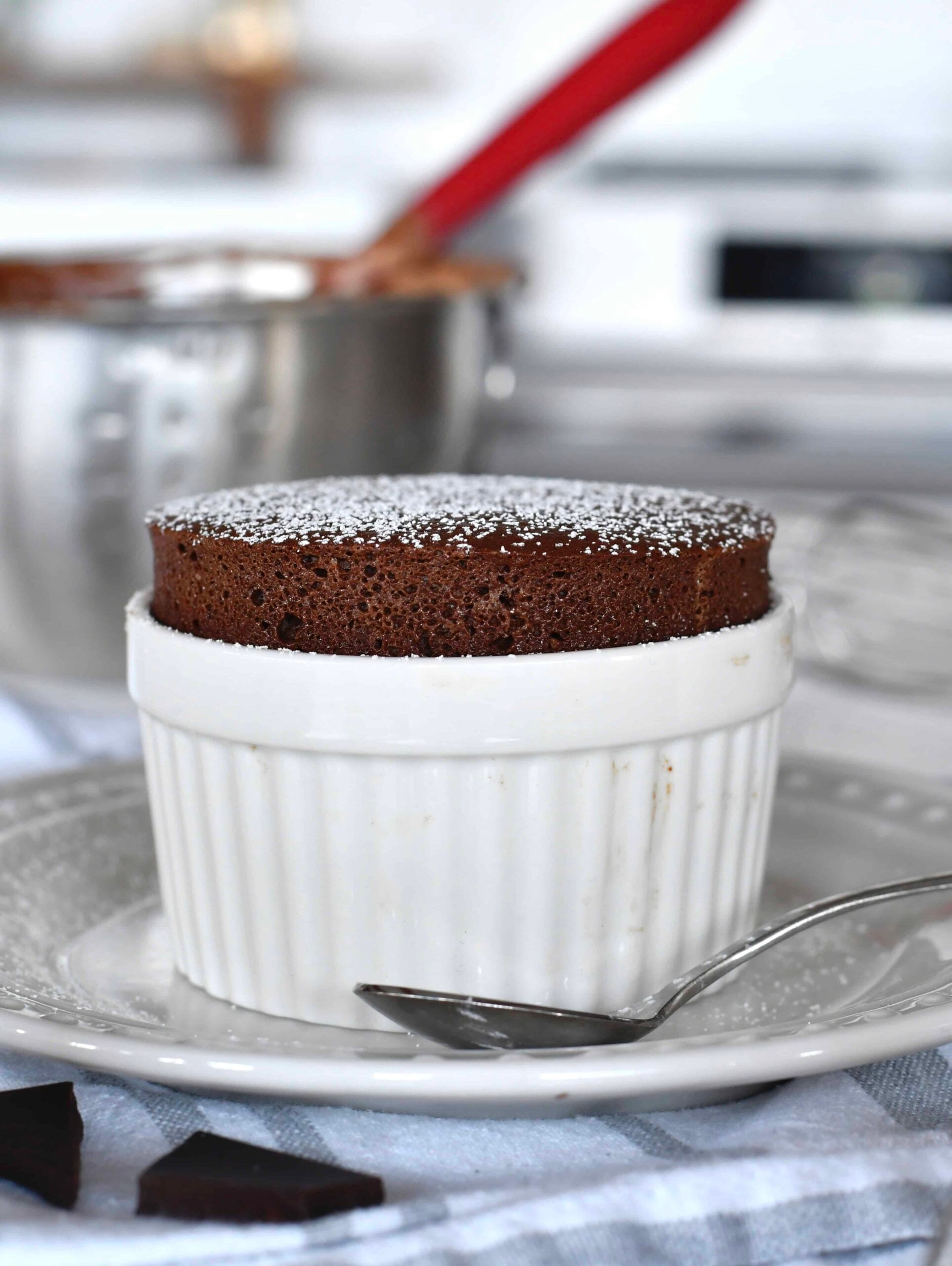 A chocolate soufflé dusted with powdered sugar is served in a white ramekin on a plate, with a spoon beside it. The background shows a mixing bowl and a kitchen setting.