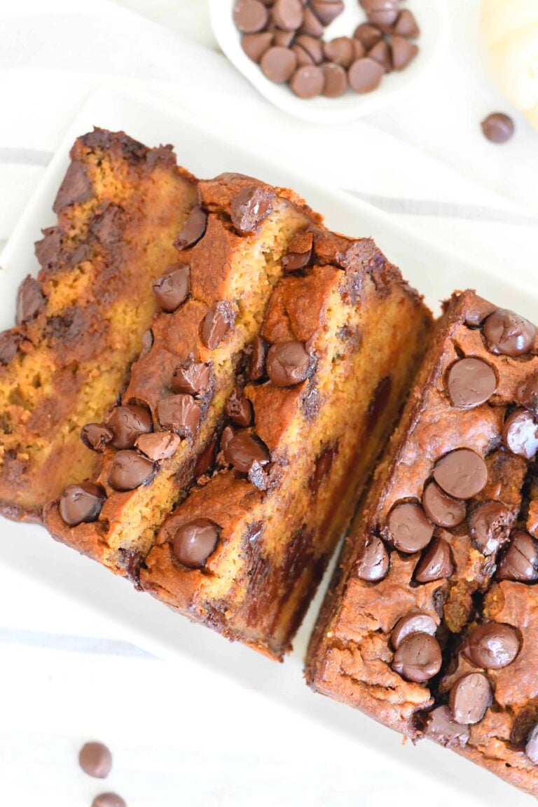 Sliced chocolate chip banana bread on a white plate, topped and filled with chocolate chips, with a small bowl of chocolate chips in the background.
