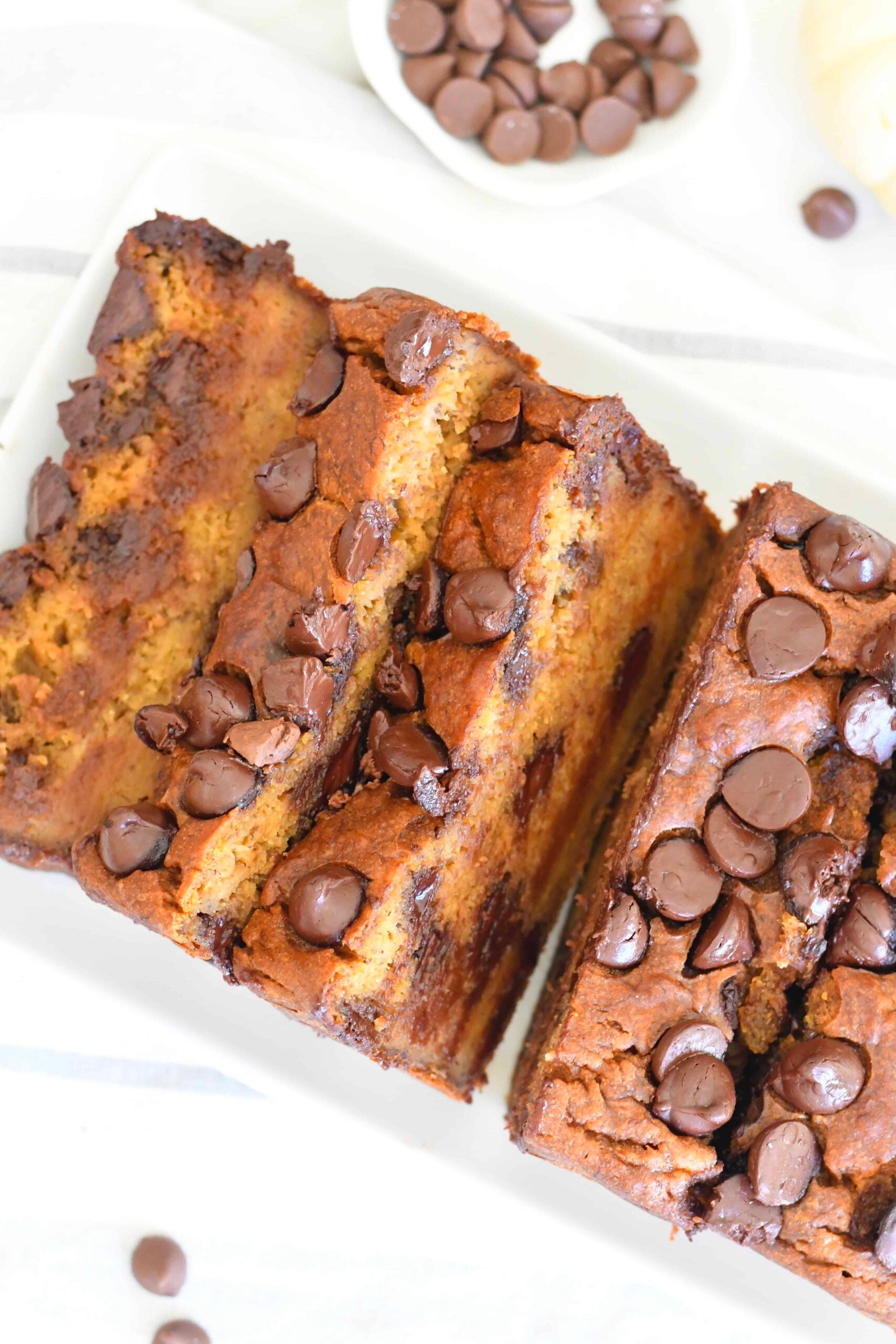 Sliced chocolate chip banana bread on a white plate, topped and filled with chocolate chips, with a small bowl of chocolate chips in the background.
