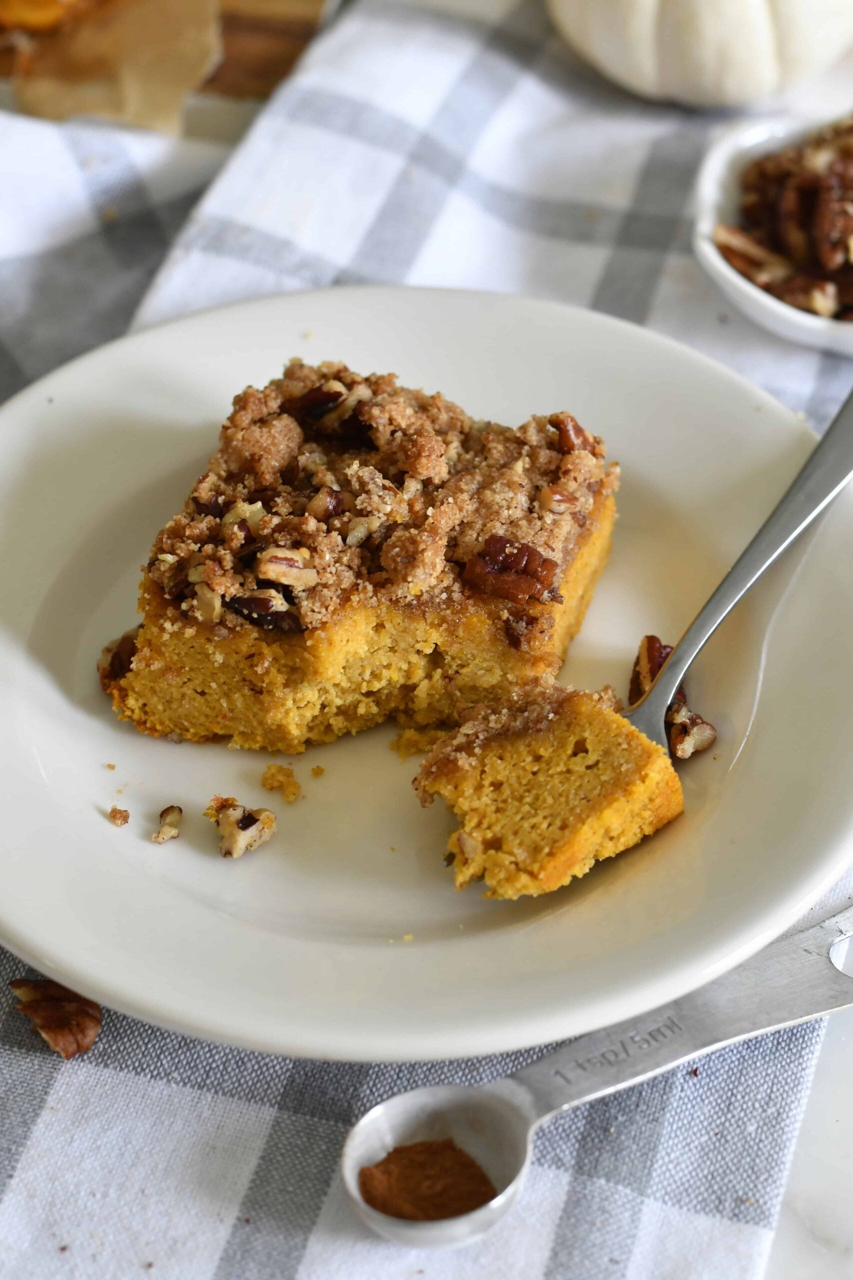 A square piece of pumpkin coffee cake with crumbly streusel topping sits on a white plate with a fork, next to scattered pecans and a spoon with cinnamon, on a gray plaid tablecloth.