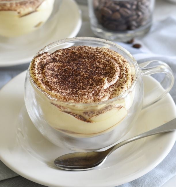 A glass cup filled with creamy tiramisu, topped with a dusting of cocoa powder, sits on a white plate with a spoon beside it. In the background, there is a glass jar of coffee beans.