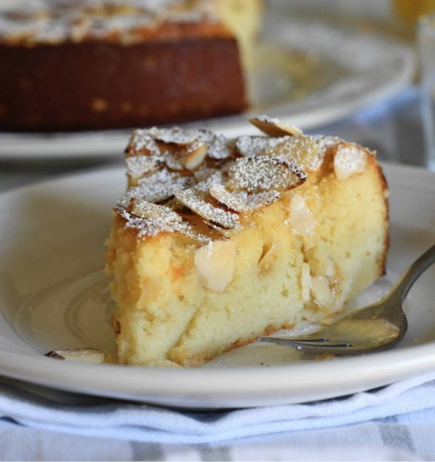 A close-up of a slice of almond cake on a white plate with a fork. The cake is topped with sliced almonds and a dusting of powdered sugar. Another cake is visible in the blurred background.