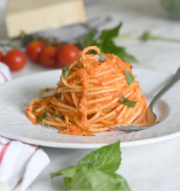 A plate of spaghetti topped with creamy tomato sauce and garnished with fresh basil leaves, with a fork twirling some pasta. Cherry tomatoes, basil, and a block of cheese are in the background.