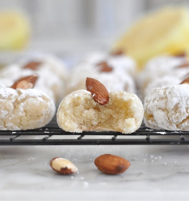 Close-up of powdered sugar-coated almond cookies on a cooling rack, with one cookie bitten to show its soft, chewy interior. Whole almonds garnish the cookies, and a few almonds rest on the marble surface.