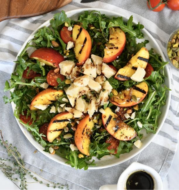 A white bowl filled with arugula, grilled peach slices, cherry tomatoes, shaved cheese, and sprinkled pistachios, drizzled with balsamic glaze, sits on a striped cloth.