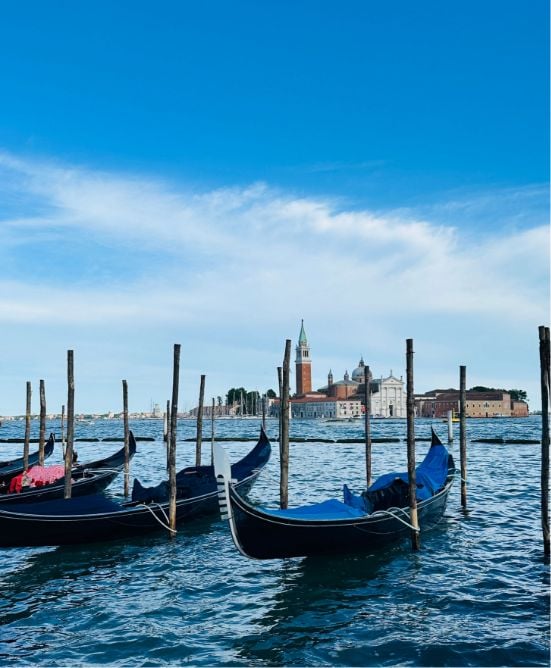 Gondolas moored on a calm canal in Venice, Italy, with wooden poles in the water and historic buildings, including a church with a tall bell tower, visible in the background under a blue sky.