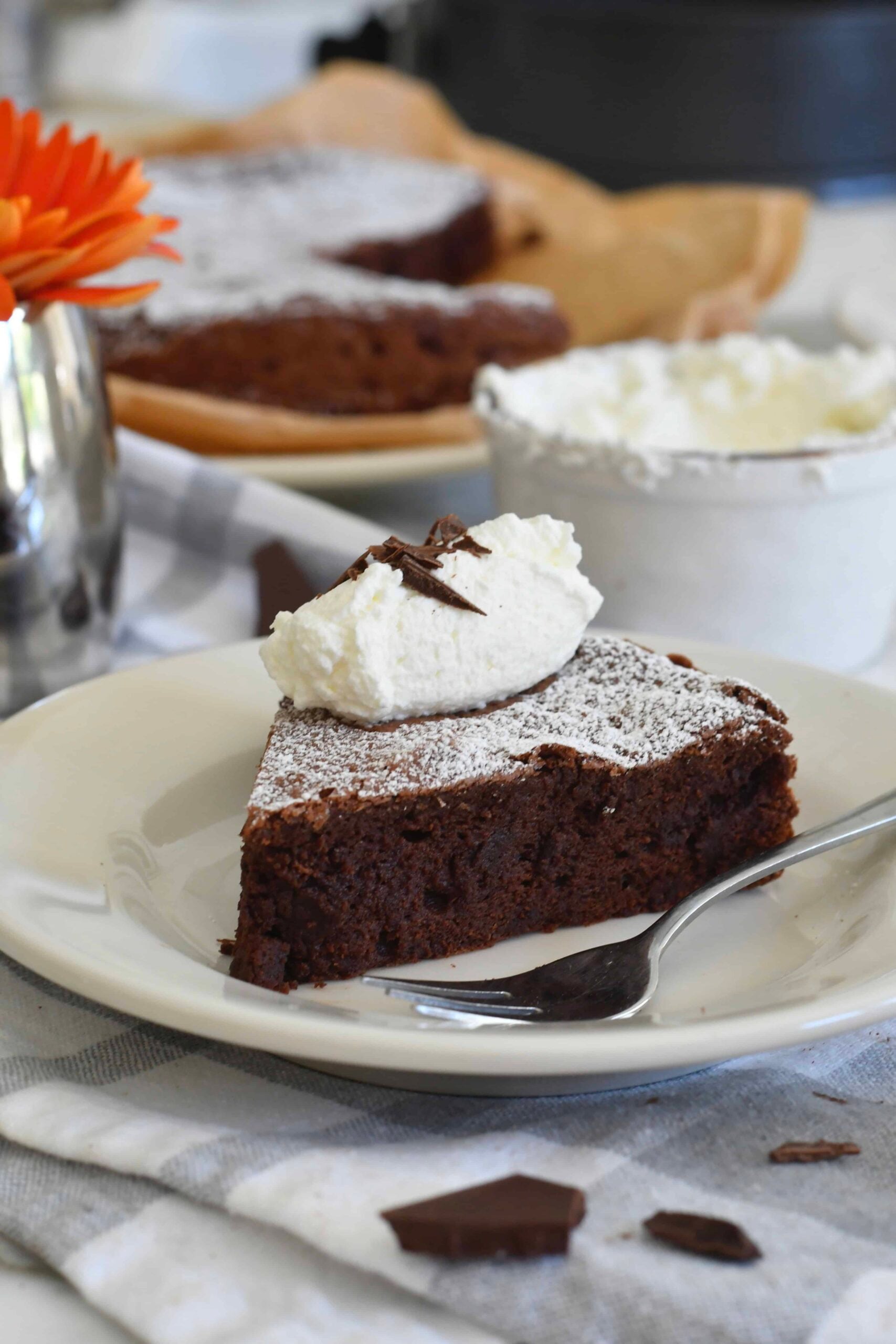 A slice of chocolate cake dusted with powdered sugar, topped with whipped cream and chocolate shavings, sits on a white plate with a spoon. A bowl of cream and another cake slice are in the background.