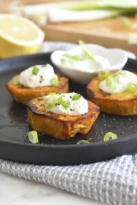 Three roasted sweet potato rounds topped with sour cream and chopped green onions are arranged on a black plate, with a bowl of sour cream and lemon in the background.
