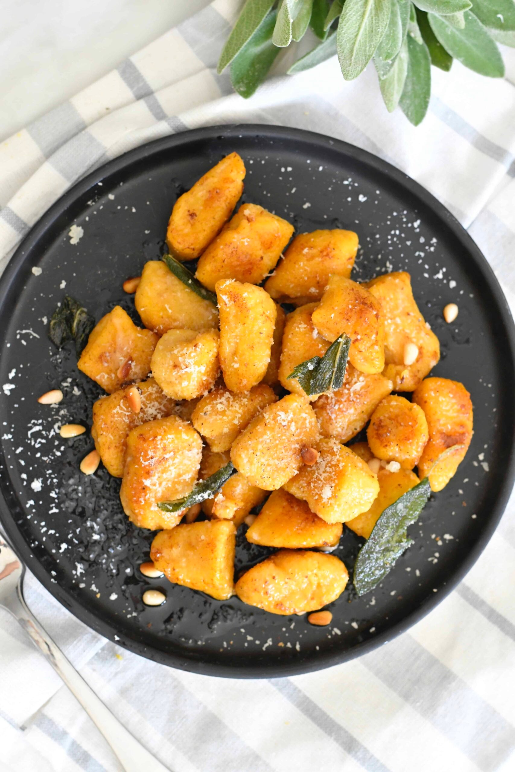 A black plate with golden brown gnocchi, garnished with grated cheese, crispy sage leaves, and pine nuts, sits on a checkered cloth beside a sprig of fresh sage.