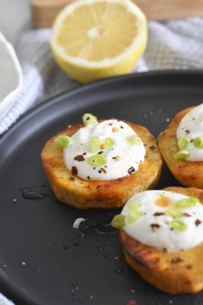 Three roasted sweet potato rounds topped with creamy white sauce, chopped green onions, and red pepper flakes are arranged on a black plate. A halved lemon is visible in the background.